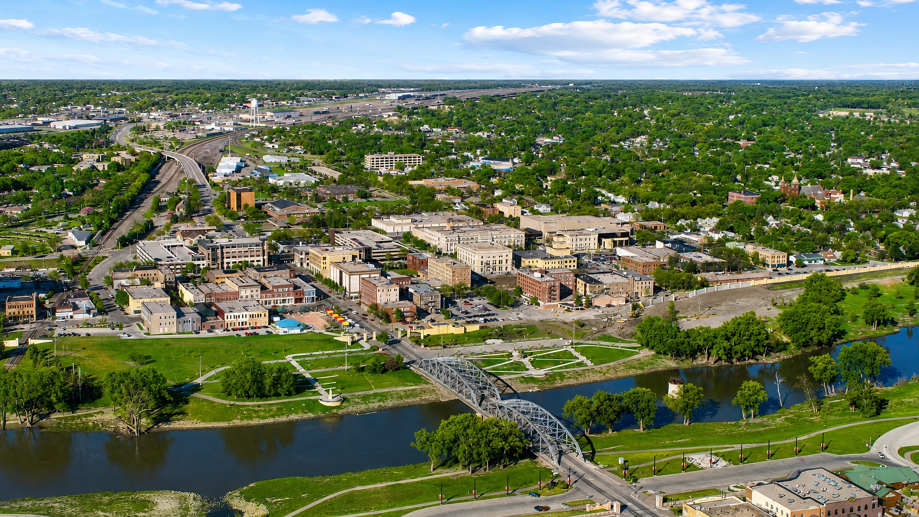 Aerial view of downtown Grand Forks, North Dakota, with its steel arch bridge over the Red River and riverside greenway.