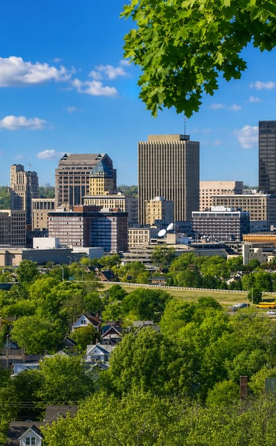Downtown Dayton, Ohio skyline of high-rise towers framed by green trees under a blue sky.