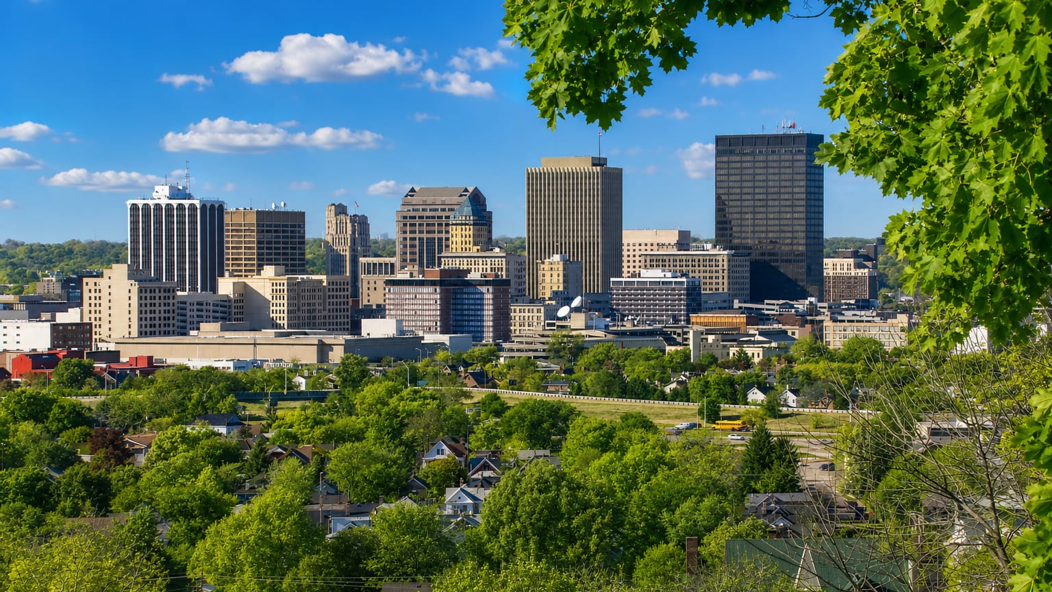 Downtown Dayton, Ohio skyline of high-rise towers framed by green trees under a blue sky.