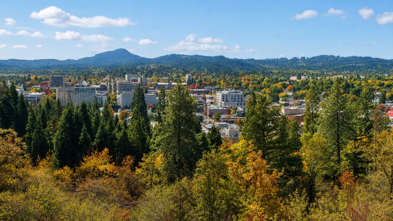 Downtown Eugene, Oregon framed by evergreen forest and autumn foliage with wooded hills behind.