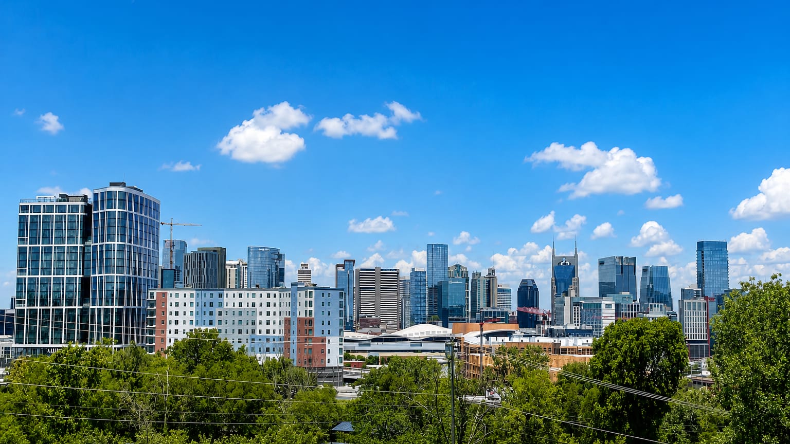 Nashville downtown mid-rise skyline seen above a line of green trees under a blue sky