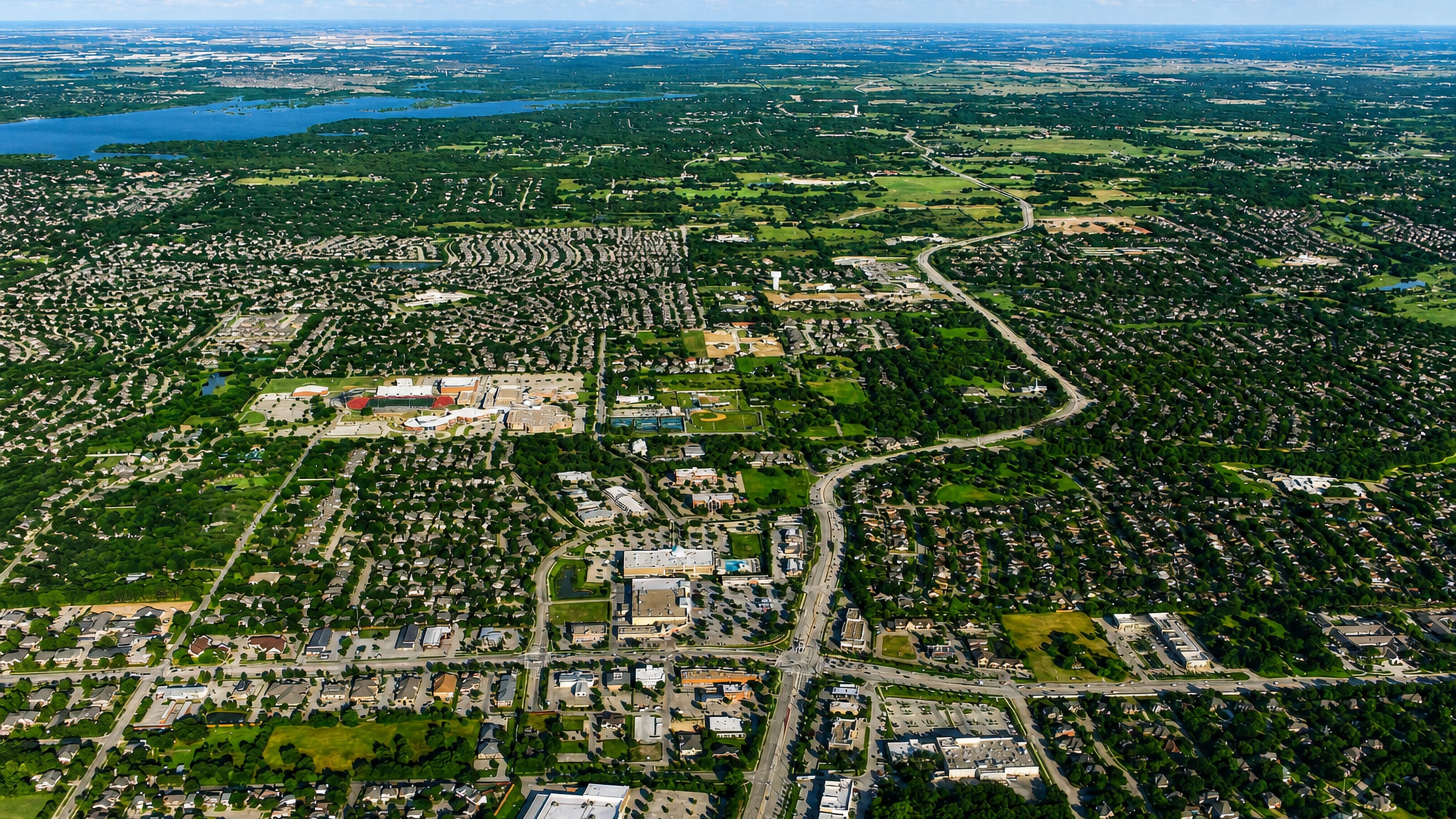 High aerial over Flower Mound, Texas, showing wooded suburbs, shopping centres and a lake beyond.