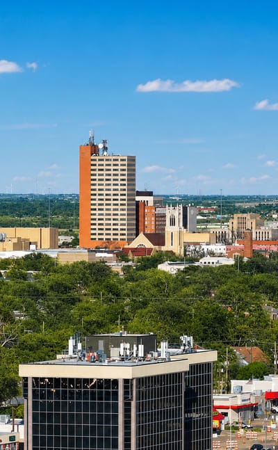 Downtown Lubbock, Texas skyline of low towers rising above green trees and the flat plains under a blue sky.