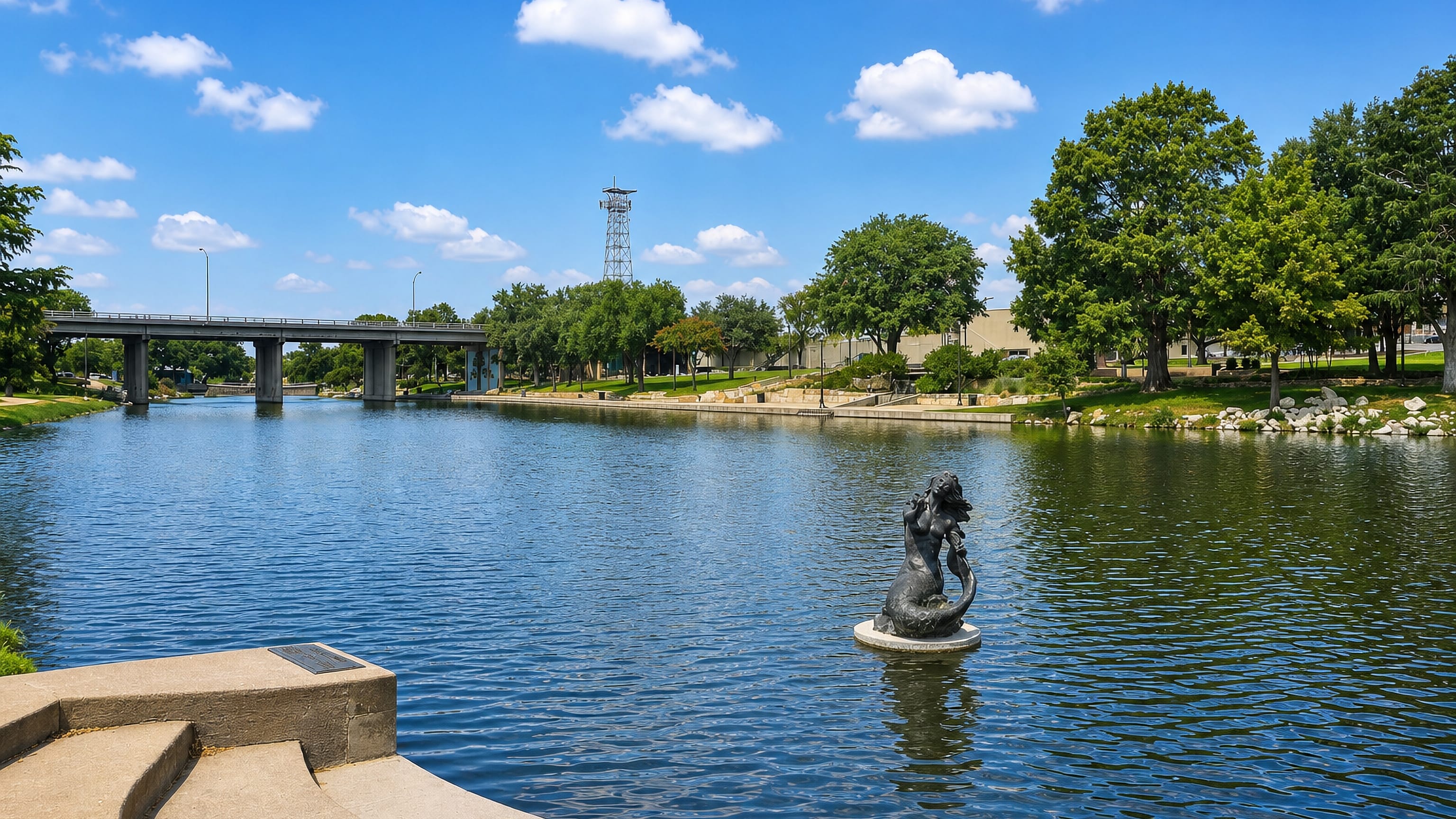 The bronze mermaid statue standing in the Concho River beside San Angelo's riverside park and bridge.
