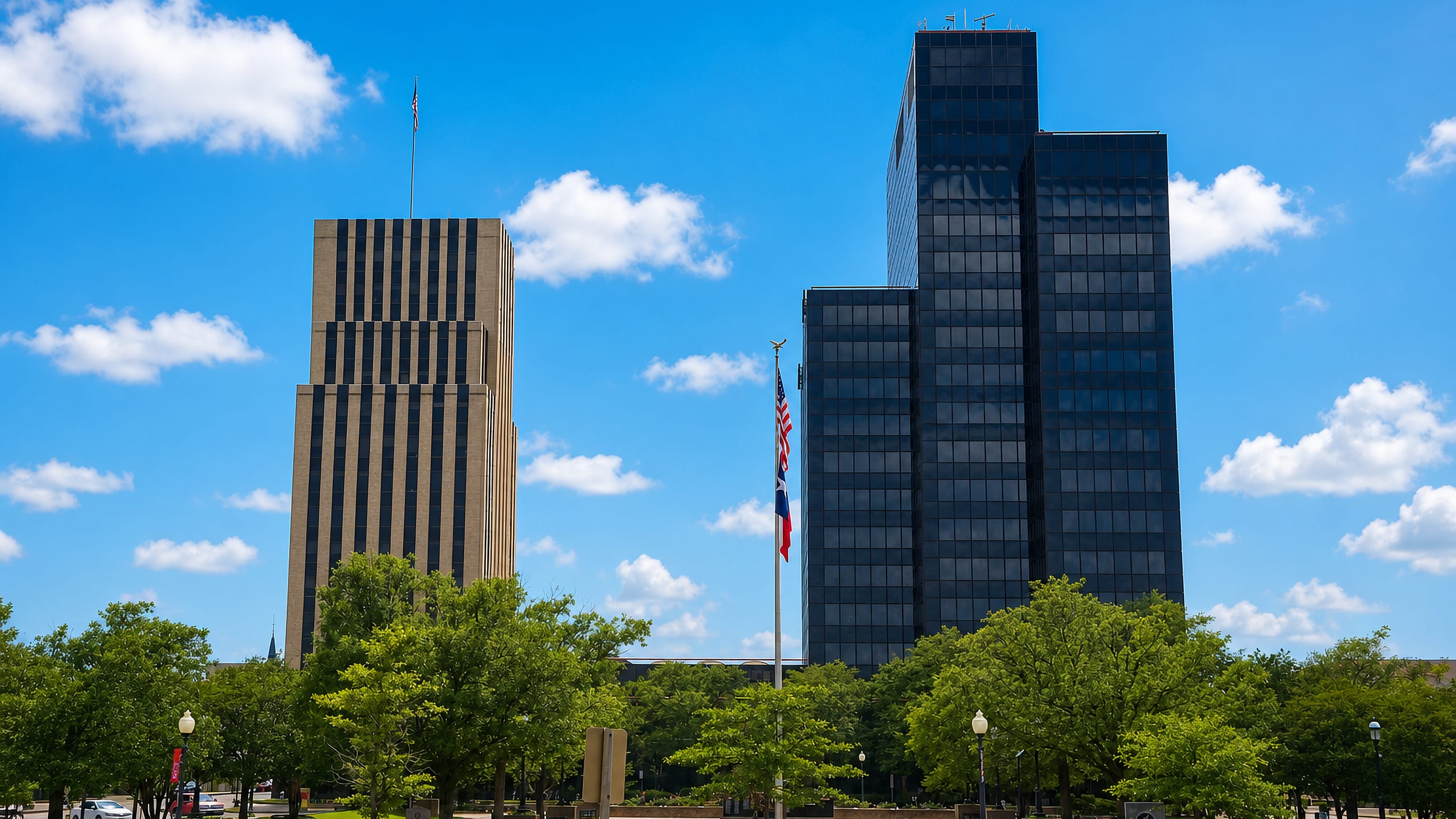 Downtown Tyler, Texas, where a dark glass office tower and a stone highrise rise above the tree-lined civic plaza.