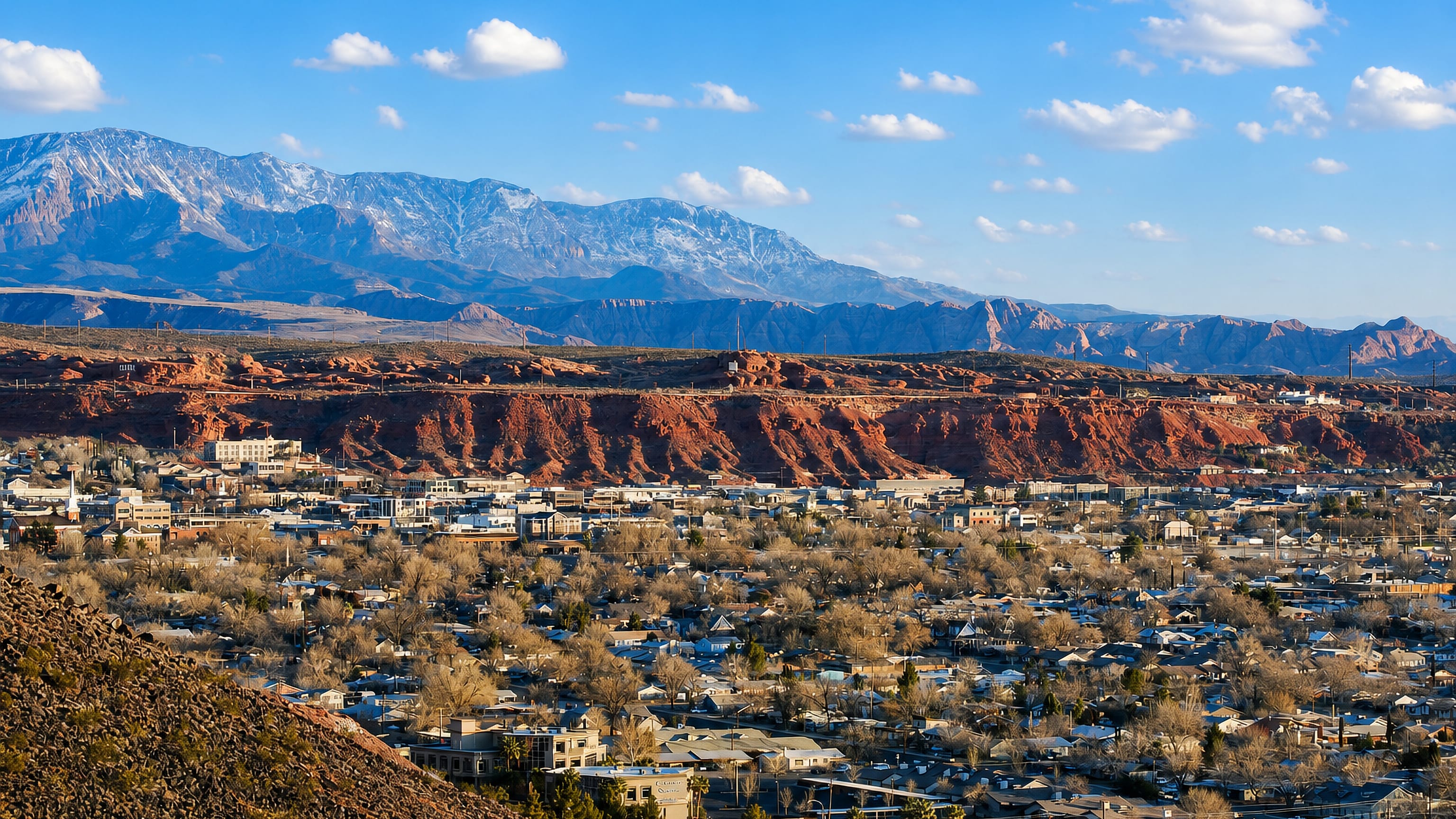 Saint George, Utah spreads below red sandstone cliffs with the snow-capped Pine Valley Mountains behind.