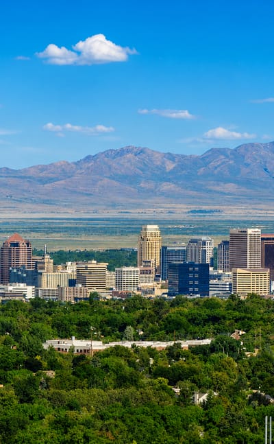 Downtown Salt Lake City skyline backed by the Wasatch Mountains, with green treetops in the foreground.