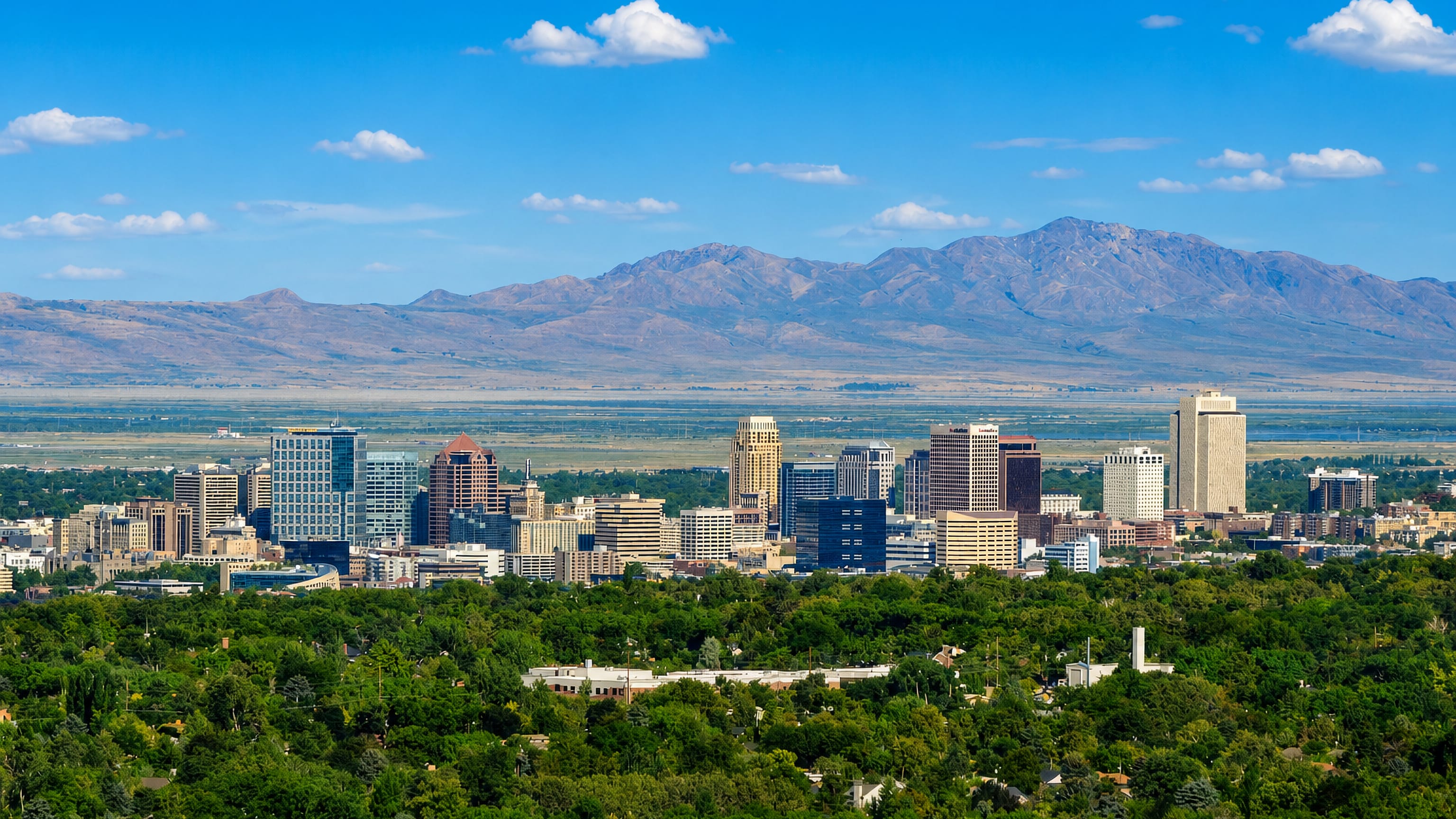 Downtown Salt Lake City skyline backed by the Wasatch Mountains, with green treetops in the foreground.