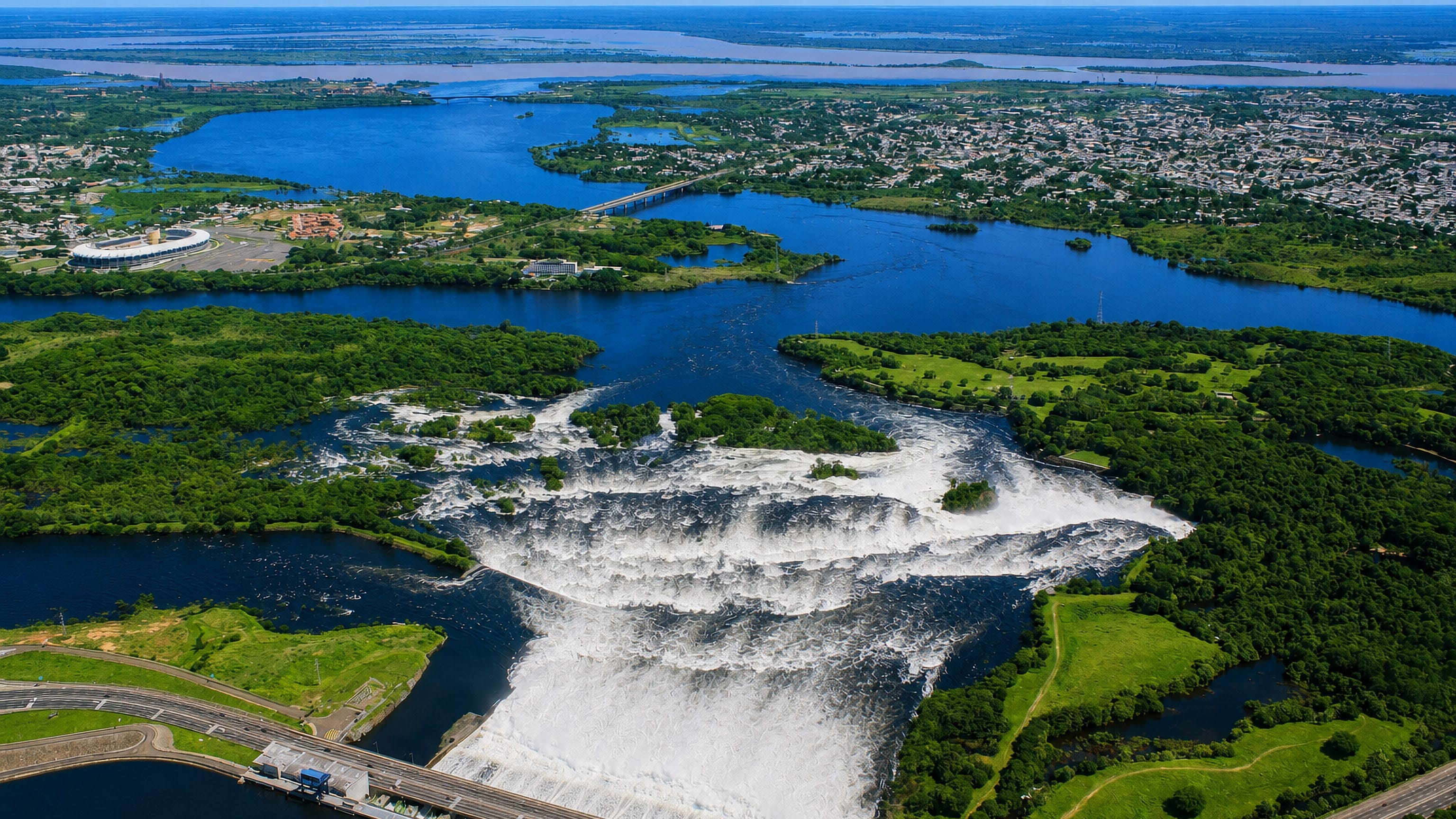 Aerial view of San Félix, Ciudad Guayana, where wide Caroní river rapids and a dam meet the city's green banks.