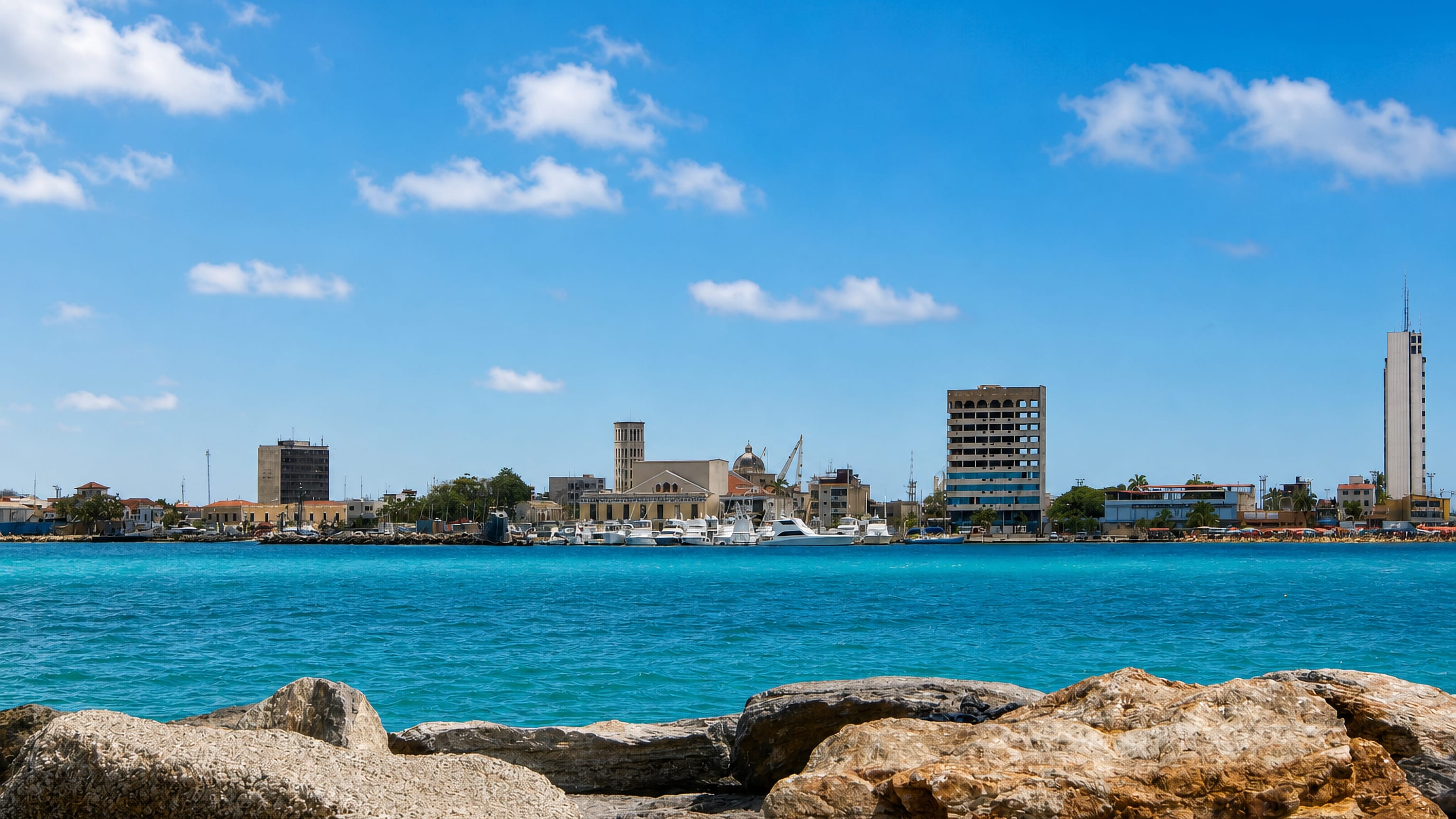 Puerto Cabello's low waterfront, church tower and marina seen across turquoise Caribbean water from a rock breakwater.