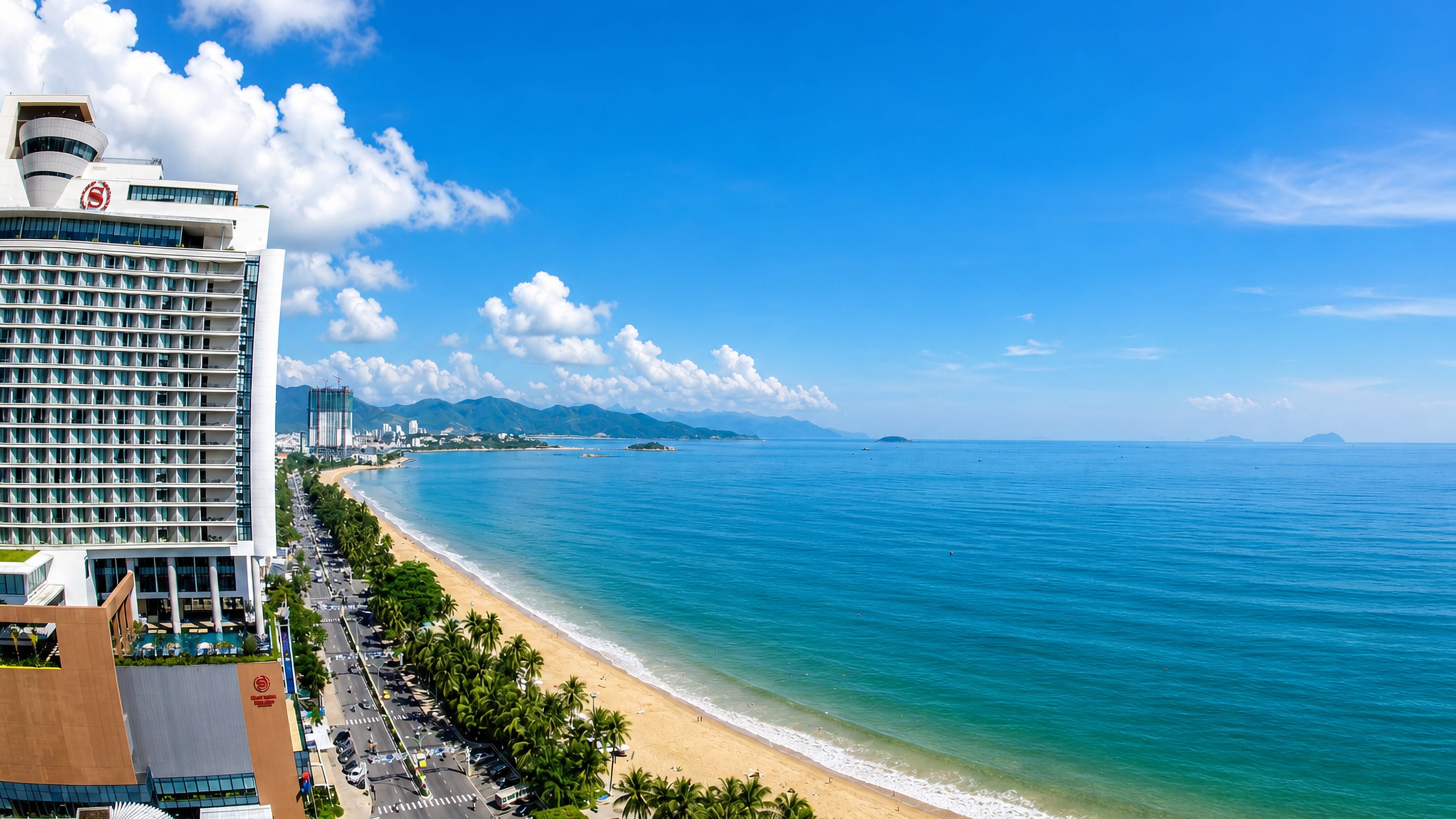 Palm-lined Tran Phu beachfront curving along Nha Trang bay past hotels toward distant mountains.