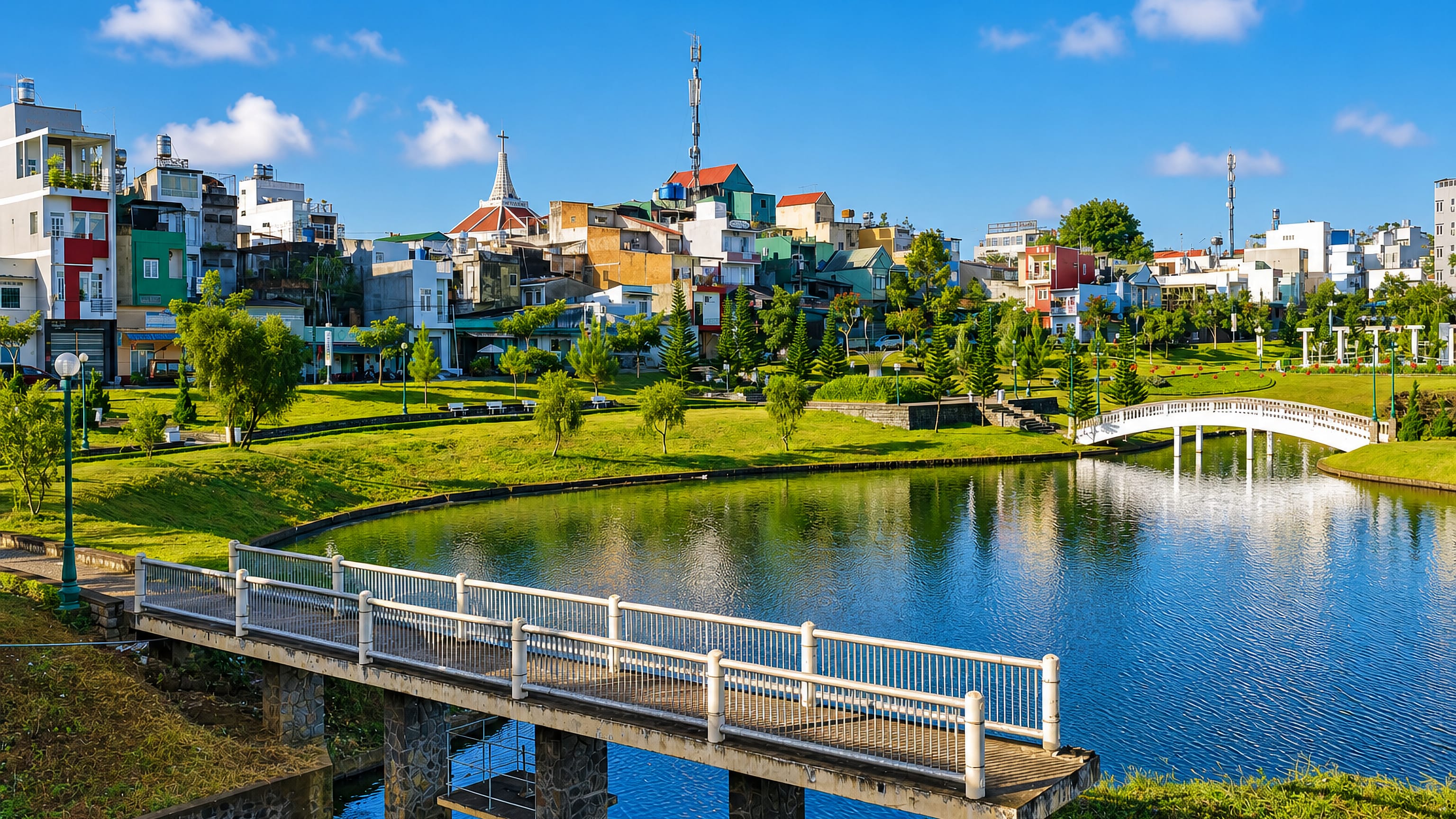 Bao Loc's hillside houses and church spire above a lakeside park and white footbridge, Lam Dong.