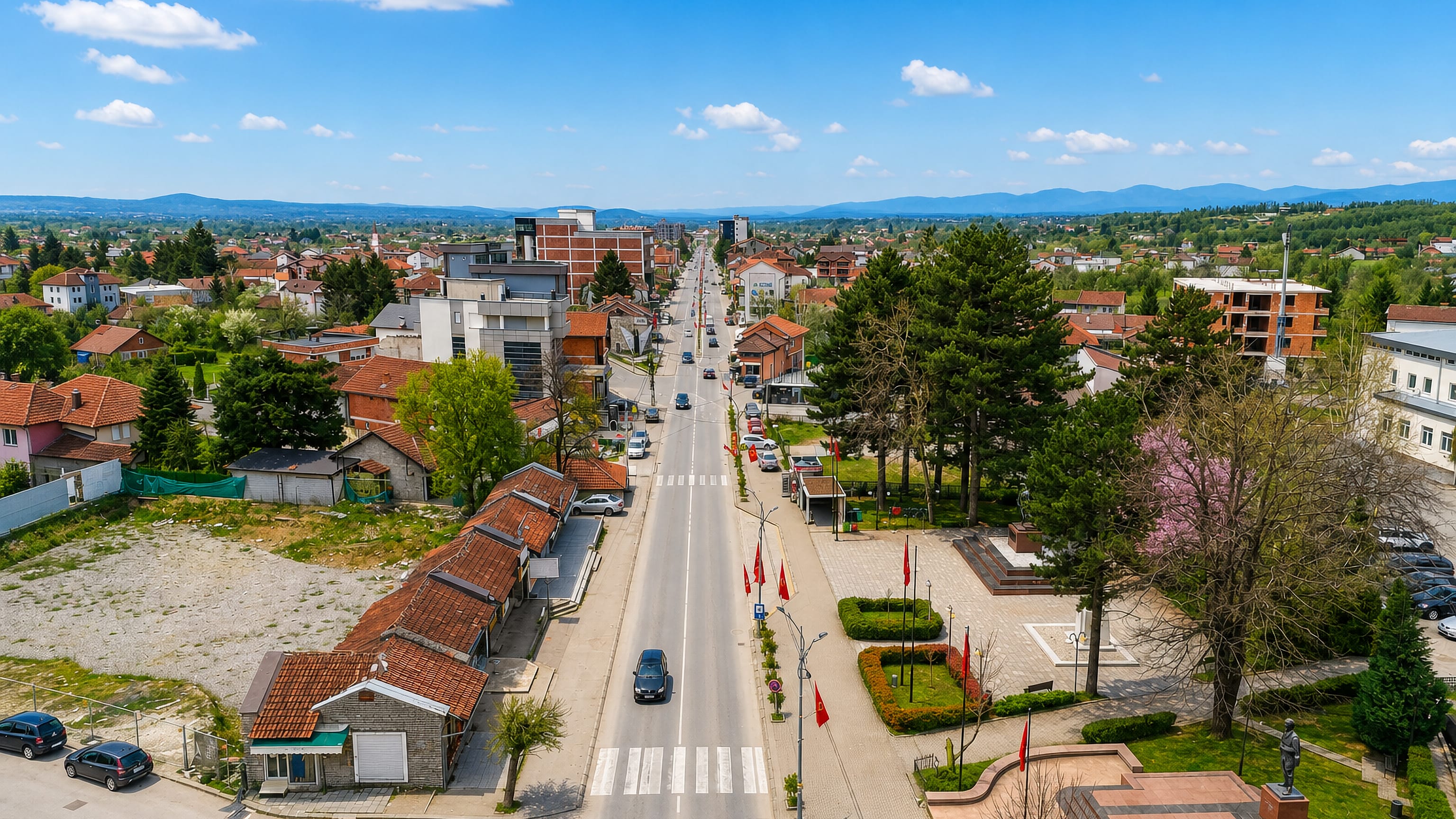 Aerial view along the main street of Deçan, Kosovo, with Albanian flags, a memorial and red-roofed houses