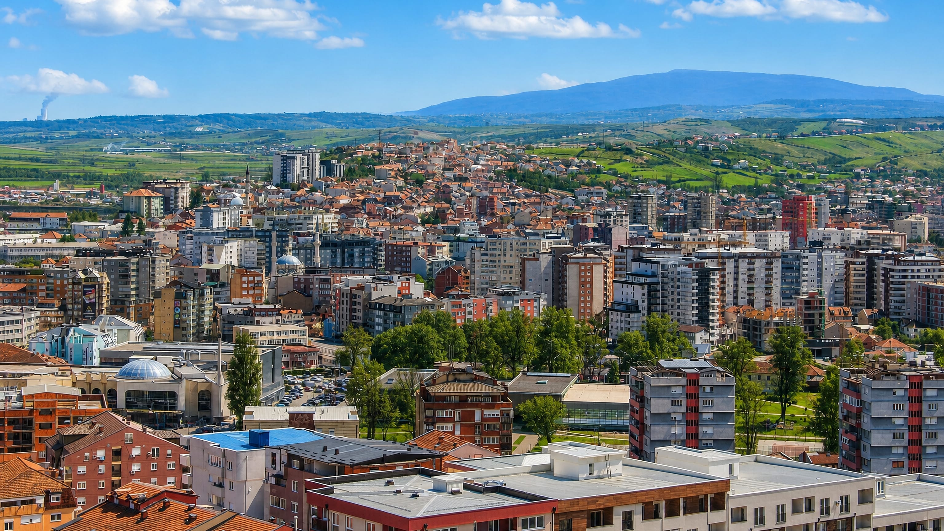 Hillside townscape of Mitrovicë, Kosovo, with mosque domes, red-tiled roofs and apartment blocks below green hills