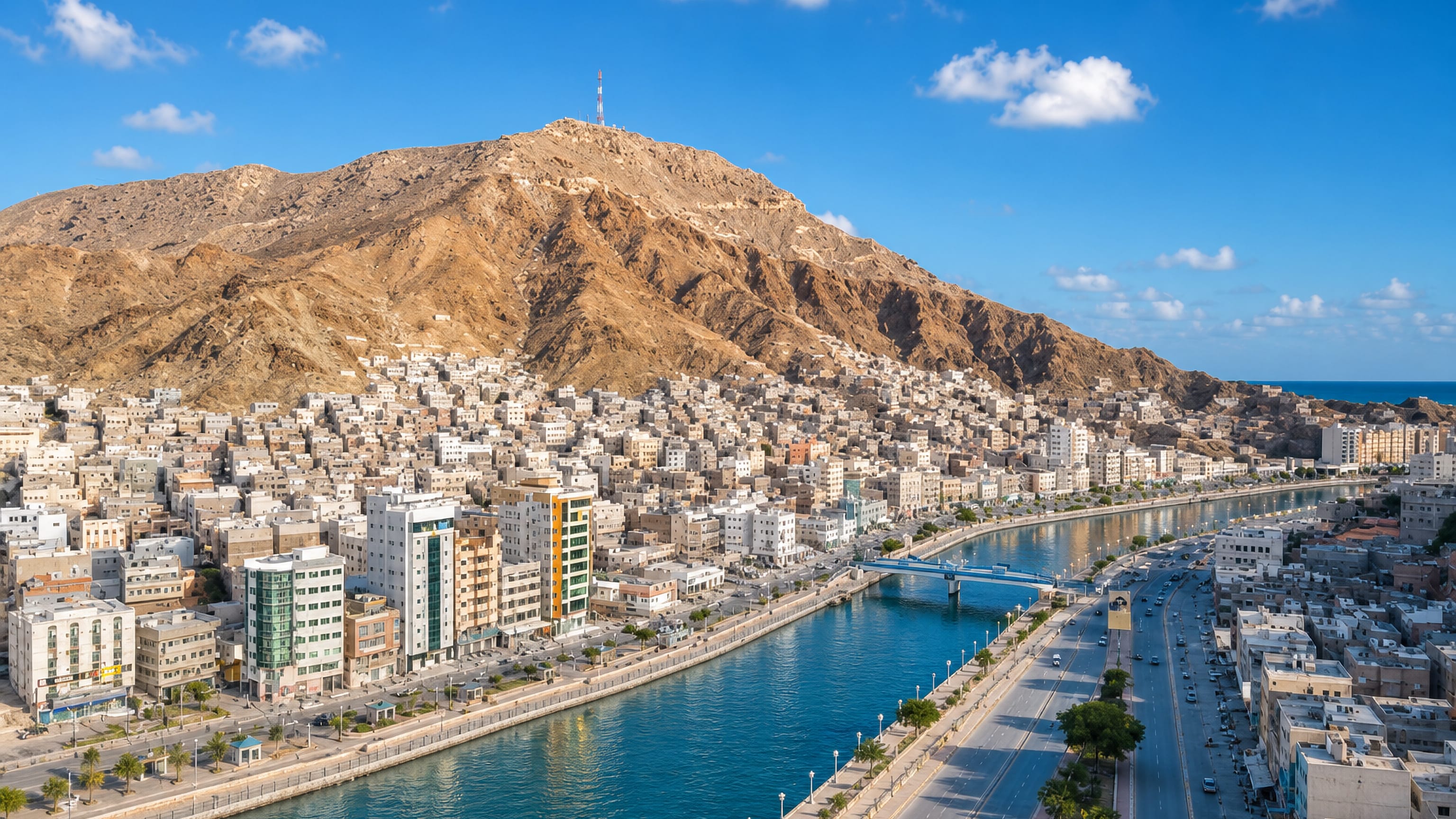 Mukalla's corniche and tidal channel in Yemen, lined with white apartment blocks below a bare coastal mountain.