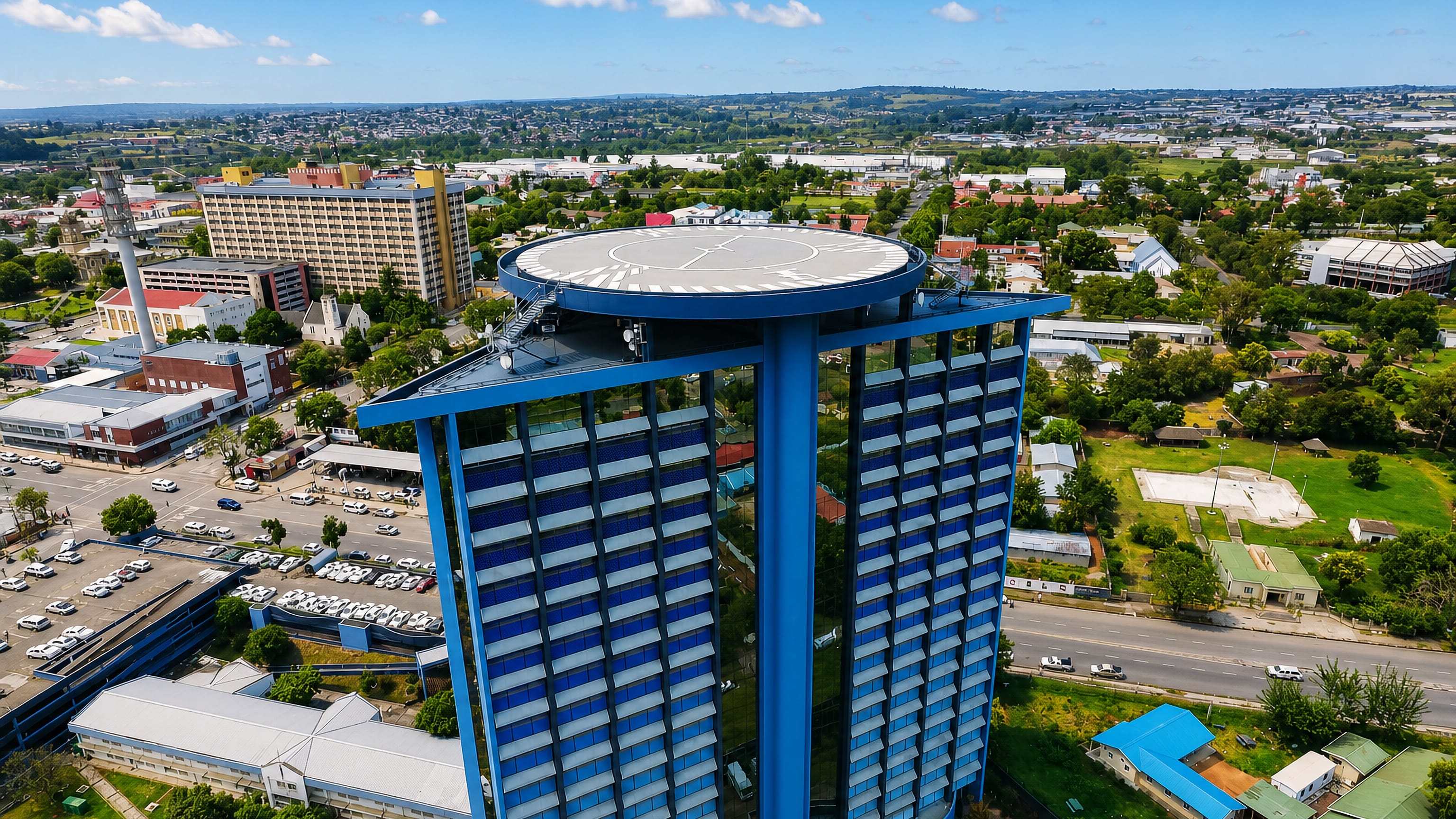 A blue glass tower with a rooftop helipad stands above the low-rise streets and green hills of Mthatha, South Africa.
