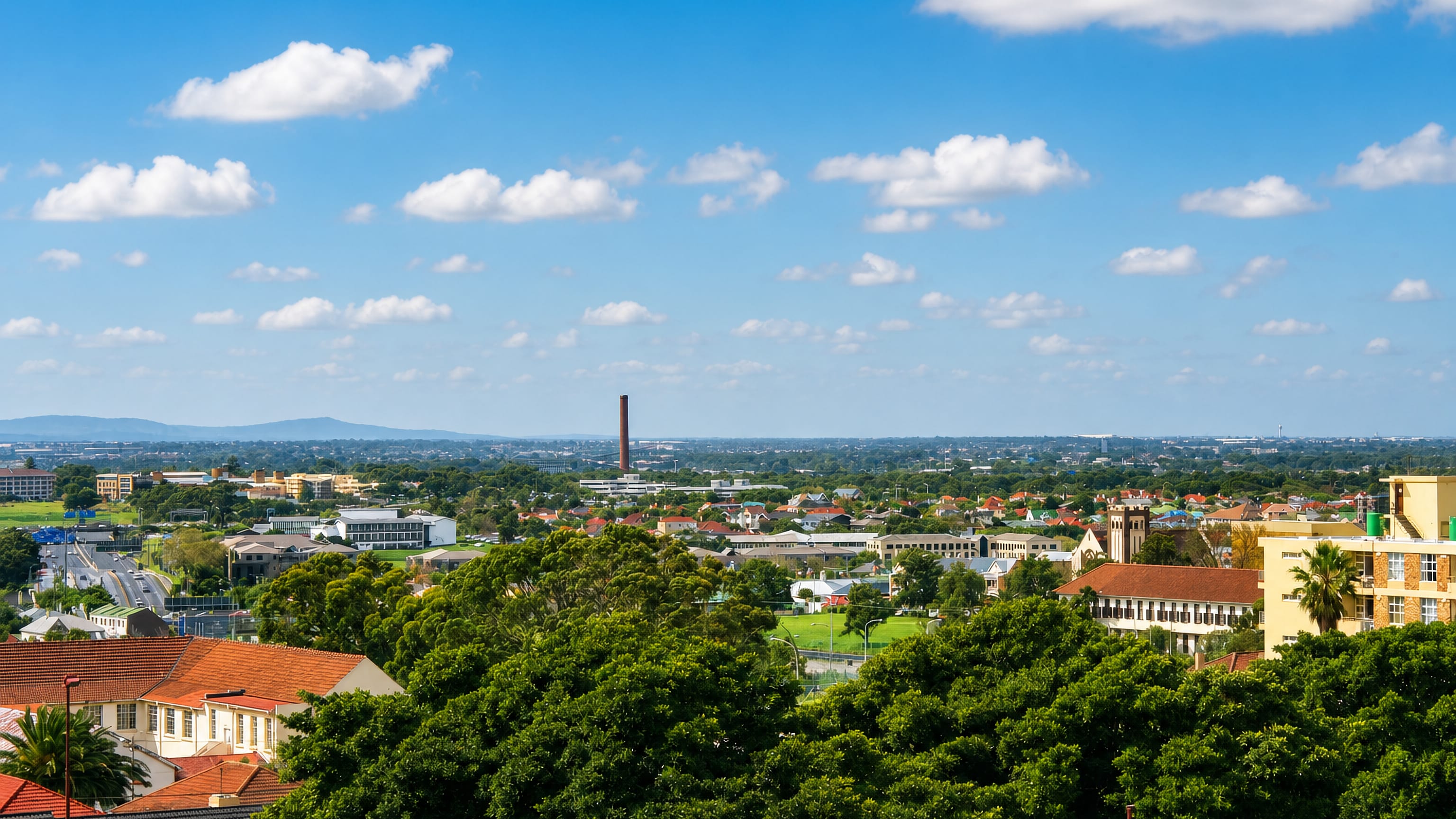 Rooftops and tree canopy of Athlone in Cape Town, with a tall brick chimney and distant mountains beyond.